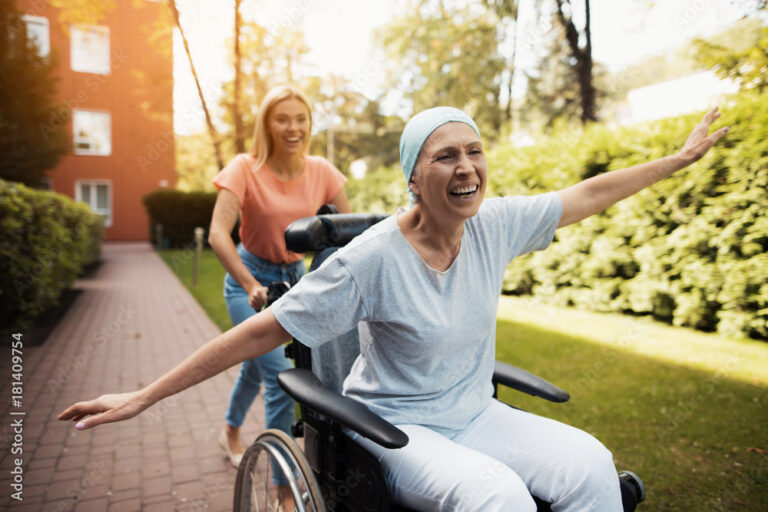 A woman with cancer is sitting in a wheelchair. She walks on the street with her daughter and they fool around.